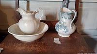 Set including a white scalloped pitcher on matching basin and floral patterned pitcher on wooden surface
