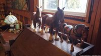Photo showing side view of three toy horses on a wooden surface near a window, two wooden, one plastic with tack.