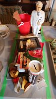Full overview of the lot showing Colonel Sanders piggy bank, red Aster cash register, and small items arranged on table.