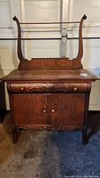 Front view of wooden wash stand showing drying rod, drawer, and cabinet doors with round knobs. Heavy, dark wood with visible grain and curved arms supporting drying rod.