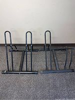 Photo of two black metal bike racks with tubular frame design, placed on carpeted floor against a wall.