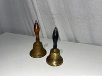 Two brass hand bells with wooden handles of different sizes captured against a white surface and backdrop.