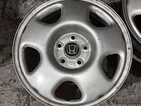 Close-up photo of one silver alloy Honda rim showing 5 lug holes and Honda emblem in center cap, with minor scratches and scuffs visible.
