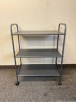 Front view of three-shelf rolling storage cart against a wall showing metal frame and caster wheels.