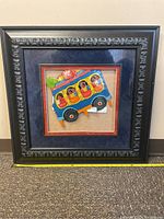 Framed South American wooden bus/shuttle art piece with ornate black wood frame and blue/orange matting, showing vibrant painted bus with passengers and goods.