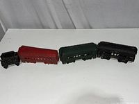 Four connected vintage cast iron train cars displayed on a white surface against a white curtain background, showing locomotive and passenger cars in black, red, and green paint.