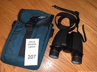 Photo showing the green fabric carrying case with buckle next to the binoculars with attached black strap, displayed on wooden floor