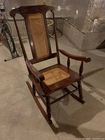 Front angled view of the wooden rocking chair showing cane seat and back, carved top rail, and armrests.