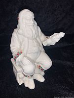 Front-left angle of white porcelain Santa figurine seated, showing colorful jeweled patterns on cuffs and boots.