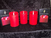Three burgundy flameless candles with two in branded boxes on black background.