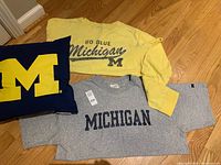 Three Michigan fan gear items on wooden floor: yellow shirt, grey sweatshirt, navy pillow with yellow M