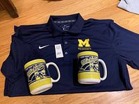 Navy blue Nike Dri-Fit polo shirt with Michigan Wolverines embroidered logo on chest, accompanied by two large Michigan coffee mugs with team graphics.
