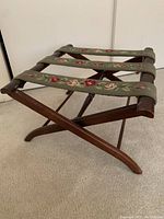 Front side angle view of the wooden luggage rack showing folded legs and needlepoint straps with floral design.