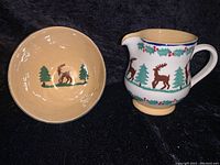 Photo of Nicholas Mosse pottery bowl and pitcher set, showing exterior handpainted deer, evergreen trees, and holly berry motifs on cream background, with bowl interior showing tan glaze and motif.