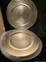 Two silver-toned charger plates shown, one resting on top of the other with visible surface texture and brushed finish.