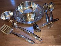 Photo showing 7 shiny silver plate and stainless steel kitchen serving items on wooden floor.