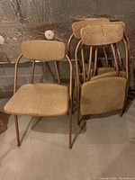 Four vintage Costco Hamilton folding chairs stacked and arranged in a basement setting, showing beige fabric upholstery and bronze metal frames.