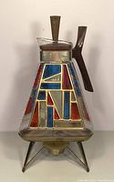Front view of vintage Inland Glass triangle carafe showing stained glass-like panels in red and blue with gold outlines, plastic handle attached to brass band, and clear textured glass lid.