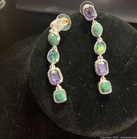 Photo showing a pair of dangling earrings with purple amethyst and green stones in silver-tone setting on black background.