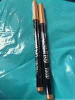 Photo of two black ABH Gel Liner eyeliners with gold caps on a blue background.