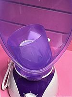 Electric facial steamer with a white base and purple detachable cone shown from a top-front angle. Power cord visible, power knob on the front.