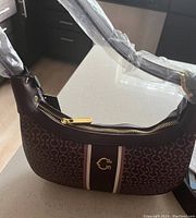 Photo showing front and side view of the brown designer purse in new condition, with protective plastic on strap.