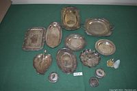 Photo showing a collection of 10 assorted silver trays in various shapes including rectangular, oval, leaf-shaped, round, and scalloped, plus 3 small silver lidded containers and 2 small shakers on a green background.