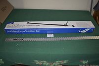 Full-length view of the HitchMate 4016 Cargo Stabilizer Bar in its box placed on a green table cloth with a ruler for scale.