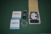 Photo showing Neurio home electricity monitor items arranged on a green surface including manuals, sensor wiring, current transformer clamp, power supply unit, and connector components.