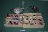 Overall view of two boxes each with a dozen divided compartments holding various vintage glass Christmas ornaments including balls in various colors, a glass teapot and candle holder nearby.