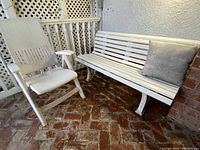 Photo showing white PVC slatted outdoor bench next to white plastic adjustable chair placed on brick floor with a pillow on the bench.