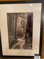 Framed sepia-tone photograph showing a winding road through tall redwood-like trees, signed and titled by the artist below the image, in black frame with white matting.