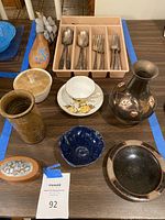 Photo of the full lot showing silverplated flatware in a plastic tray, stoneware vases, dishes, floral china cup and saucer, wooden box, and painted wooden shoe sizing tool.