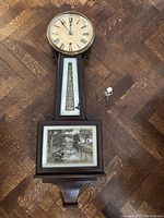 Full view of the antique New Haven banjo clock showing dial, pendulum section, lower artwork, key, and pendulum piece on wooden floor.