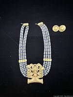 Full view of necklace with blue, green, and white beads and central gold-plated pendant, alongside matching gold-plated circular earrings on black background.