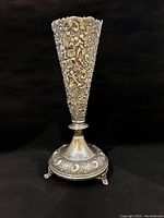 Full front view of the silver vase showing detailed embossing, flared top, and ornate base with three feet.