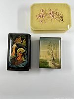 Top view of the three boxes showing decorative tops and relative sizes: alabaster floral box, black lacquer box with folk art, and painted nature scene box