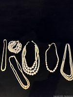 Six pearl and faux pearl jewelry pieces laid out against black background, showing variety of necklaces and bracelet.
