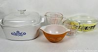 White CorningWare square casserole dish with blue cornflower pattern, two Pyrex measuring cups, orange-brown Pyrex mixing bowl, and clear glass lid arranged for display