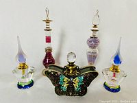 Five decorative perfume bottles displayed on a flat surface showing the enamelled metal butterfly bottle and four thin art glass bottles with various stoppers.