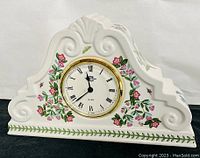 Front view of the white ceramic mantle clock decorated with pink flowers and green leaves surrounding the clock face with Roman numerals and gold trim.