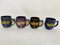 Front view of four handmade Robert Fisher pottery mugs in blue, purple, brown, and blue, each with yellow dragonfly design and comfortable handle.