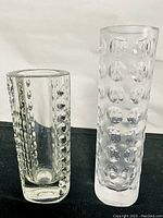 Two cylindrical glass vases side by side, one clear and one frosted, both with raised bubble patterns on a black background.