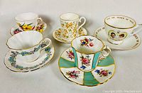 Five bone china cups and saucers displayed together, showing the colorful floral and commemorative prints and gold trim.