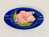 Top view of the oval ashtray showing bright pink magnolia flower with green leaves on deep blue background.