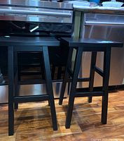 Two black wooden bar stools standing on a wooden floor in front of kitchen appliances.