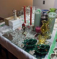 Photo showing a variety of candleholders, glassware, yellow glass oil lamps, red twisted taper candles, battery-operated candles, and a Willow Tree figurine in the background