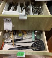 Two kitchen drawers with one holding silver flatware including forks, spoons, and knives, and the other holding various kitchen utensils including slotted spoon, dough cutter, meat tenderizer, ice cream scoop, tongs, can opener, corkscrew, tea strainers, measuring tape, and scissors