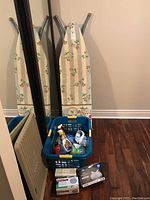 Photo showing ironing board with floral cover, blue laundry basket with cleaning supplies, gloves and masks boxes stacked on floor.