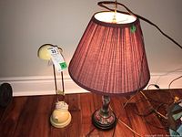 Photo shows the two lamps placed on a wooden floor, one ceramic and metal candlestick-style lamp with purple lampshade, and one smaller cream-colored plastic desk lamp with a brass arm.